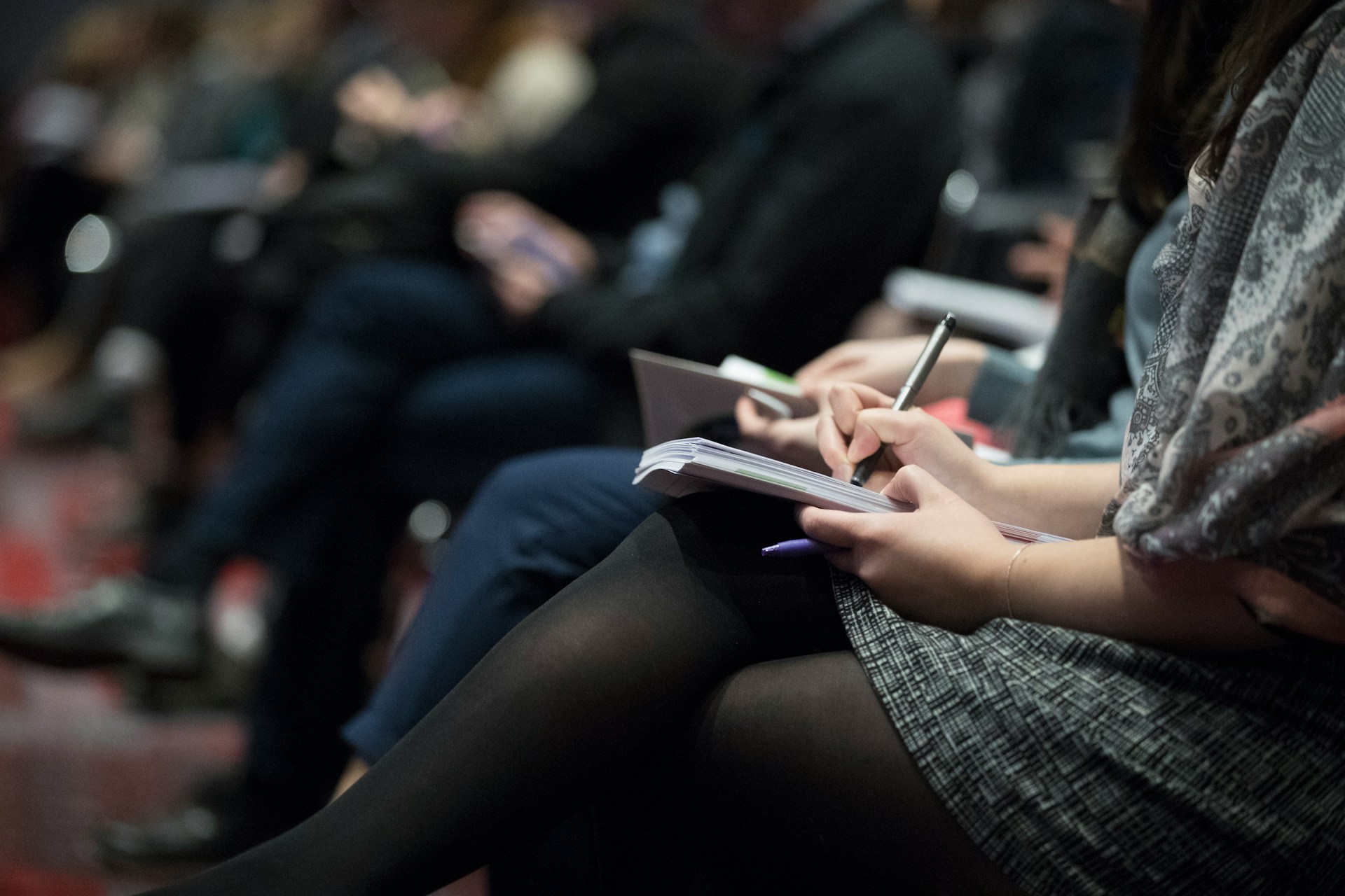 Writers sit in a row, taking notes, wearing dark clothing, only the closest writer is in focus