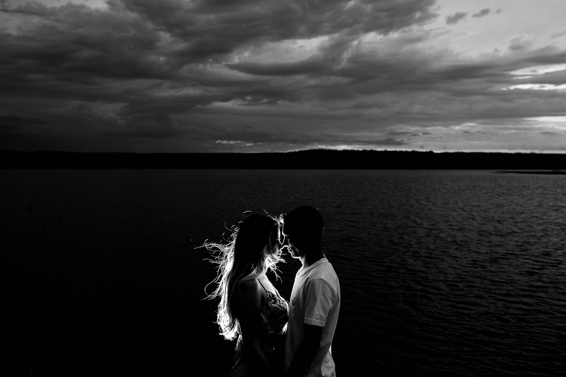 Man and woman stand close together with a large body of water in the background rendered in black and white